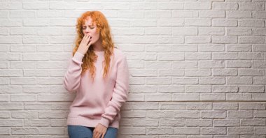 Tuğla duvardan esneme sıkılmış duran genç Kızıl saçlı kadın kapsayan ağız el ile yorgun. Restless ve uyku hali.