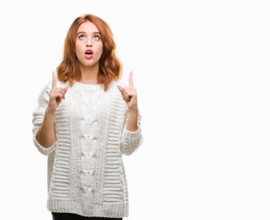 İzole arka plan kış kazak giyen genç güzel kadın hayrete ve yukarıya bakıyor ve parmakları ile işaret ve silah kaldırdı şaşırttı.