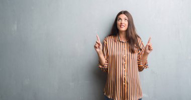 Genç esmer kadın grunge gri duvarın üzerinden hayran ve yukarıya bakıyor ve parmakları ile işaret ve silah kaldırdı şaşırttı.