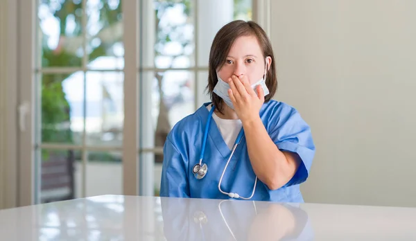 Down syndrome woman wearing nurse uniform cover mouth with hand shocked with shame for mistake, expression of fear, scared in silence, secret concept