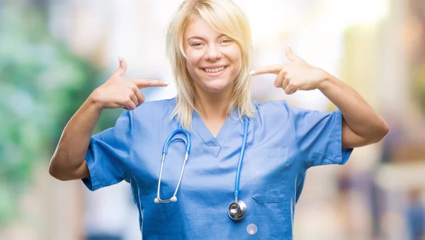 年轻漂亮的金发女医生穿着医疗制服在孤立的背景下微笑着自信地显示和指着手指的牙齿和嘴巴 健康理念 — 图库照片