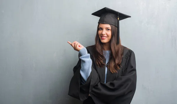 非常に幸せな側の手と指で指す大学院の制服を着てグランジ灰色の壁を越えて若いブルネットの女性 — ストック写真