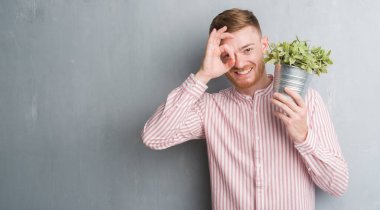 Genç Kızıl saçlı adam bitki pot ile ok işareti ile el parmaklarının arasından seyir göze yapıyor gülümseyen mutlu yüz tutan gri grunge duvarın üzerinden