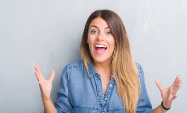 Gri grunge Genç Yetişkin kadın duvar büyük gülümseme ile çığlık zaferini kutluyor ve ellerini kaldırdı giyen denim kıyafeti çok mutlu ve heyecanlı, kazanan ifade
