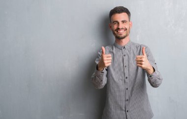 Genç yetişkin adam duvar gri grunge onaylama üzerinde ayakta el ile pozitif jest yapıyor gülümseyen ve başarı için mutlu yaşasın. Kamera, kazanan jest arıyorsunuz.