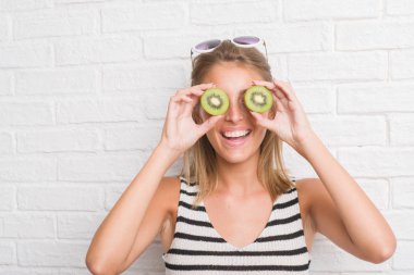 Beyaz tuğla duvar yeme üzerinde güzel genç kadın yeşil kivi ayakta ve diş gösterilen kendine güvenen bir gülümsemeyle gülümseyerek mutlu bir yüz ile