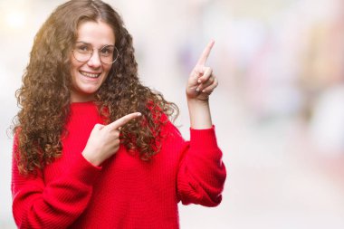 Güzel esmer kıvırcık saçlı Genç kız gülümseyerek ve iki elleri ve parmakları ile tarafına işaret kamera bakarak izole arka plan üzerinde gözlük ve kış kazak giyiyor.