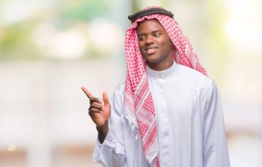 Kameraya bakarak yan el ve parmak ile işaret eden geleneksel keffiyeh yüzünde kocaman bir gülümseme ile izole arka plan üzerinde giyen genç Arapça Afrika adam.