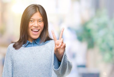 Mutlu yüz zafer işareti yaparken kameraya göz kırpıyor ile gülümseyen izole arka plan üzerinde kış kazak giyen genç Asyalı kadın. İki numaralı.