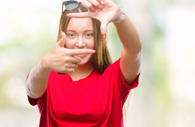 Genç güzel beyaz kadın elle yapma çerçeve ve parmakları ile mutlu yüz gülümseyen izole arka plan üzerinde güneş gözlüğü takıyor. Yaratıcılık ve Fotoğrafçılık kavramı.