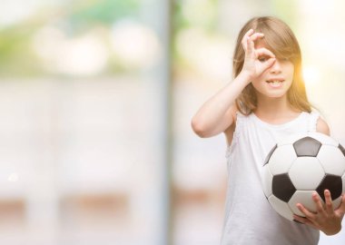 Genç Sarışın yürümeye başlayan çocuk futbol topu ile ok işareti ile el parmaklarının arasından seyir göze yapıyor gülümseyen mutlu yüz tutan