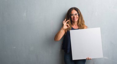 Parmak, mükemmel sembolü ile ok işareti yapıyor boş afiş tutarak gri grunge duvarın üzerinden duran orta yaş İspanyol kadın