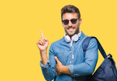 Yüzünde kocaman bir gülümseme ile izole arka plan üzerinde kulaklık ve sırt çantası giyen genç yakışıklı turist adam kameraya bakarak yan el ve parmak ile işaret.