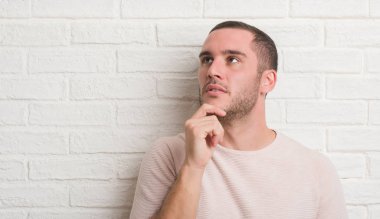 Genç beyaz adam beyaz tuğla duvar ciddi bir yüz düşünme hakkında soru, çok karışık bir fikir üzerinde ayakta