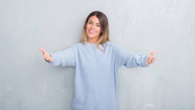 Gri grunge duvar sarılmak için kollarını açarak gülümseyerek kameraya bakarak kış kıyafeti giymiş genç yetişkin kadın. Neşeli ifade kucaklayan mutluluk.