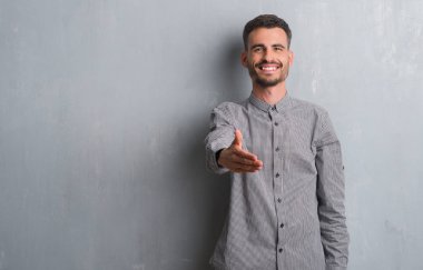 Tebrik ve konuksever dost sunan karşılıklı gülümseyen gri grunge duvarın üzerinden ayakta genç yetişkin adam. Başarılı iş.