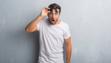 Yakışıklı genç adam korkuyor ve şok sürpriz ifade, korku ve heyecan yüz ile güneş gözlüğü takıyor gri grunge duvarın üzerinden.