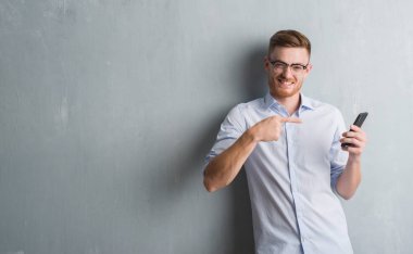 Genç Kızıl saçlı adam gri grunge duvar el ve parmak ile işaret çok mutlu smartphone kullanarak ileti gönderme