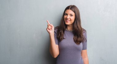 Grunge genç esmer kadın duvar bir fikirle sürpriz gri veya gülen yüz, bir numara ile işaret parmağı soru