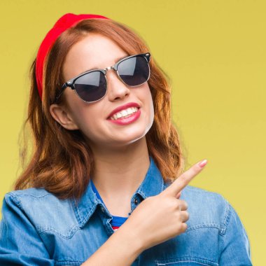 Gülümseyen ve iki elleri ve parmakları ile tarafına işaret kamera bakarak güneş gözlüğü takıyor izole arka plan üzerinde genç güzel kadın.