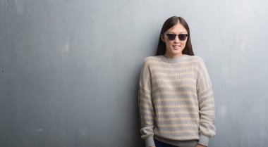 Genç Çinli kadın ayakta ve diş gösterilen kendine güvenen bir gülümsemeyle gülümseyerek mutlu bir yüz ile güneş gözlüğü takıyor gri duvarın üzerinden