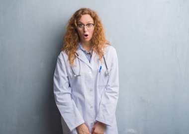 Genç redhead doktor kadın korkuyor ve sürpriz ifade, korku ve heyecan yüz ile şok bir palto giyiyor gri grunge duvarın üzerinden.