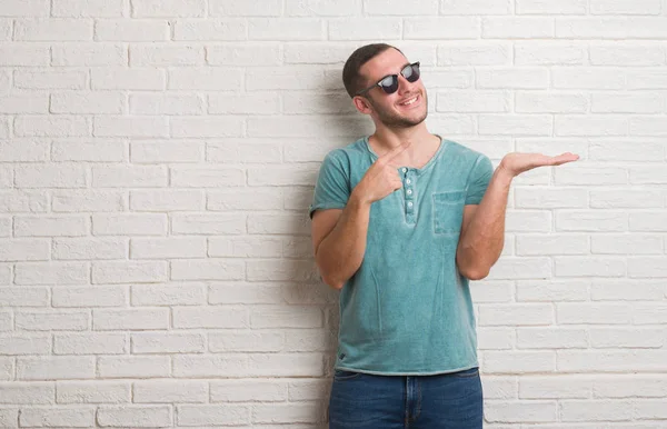 El ve parmak ile işaret çok mutlu güneş gözlüğü takmış beyaz tuğla duvarın üzerinden ayakta beyaz delikanlı