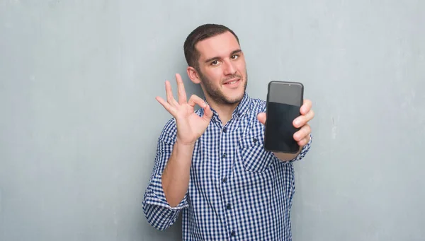 年轻的白种人在灰色的垃圾墙显示屏幕的智能手机做 符号与手指 优秀的符号 — 图库照片