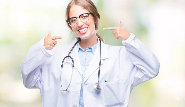 美丽的年轻金发碧眼的医生妇女穿着医疗制服在孤立的背景微笑自信显示和指向与手指牙齿和嘴 健康理念 — 图库照片