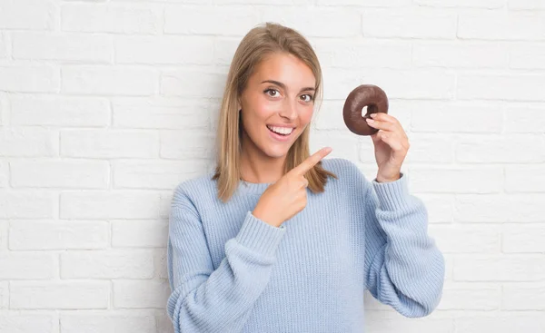 チョコレート ドーナツの手と指で指している非常に幸せを食べる白いレンガの壁に美しい若い女性 — ストック写真