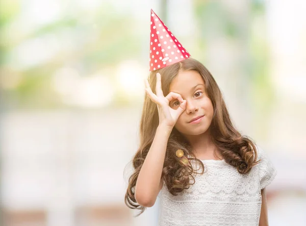 指の間から見て目に手で サインをして笑っている幸せそうな顔の誕生日の帽子をかぶってヒスパニックのブルネットの少女 — ストック写真