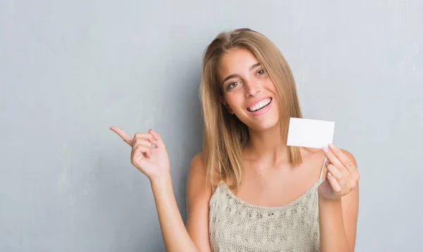 Beautiful young woman over grunge grey wall holding blank visit card very happy pointing with hand and finger to the side