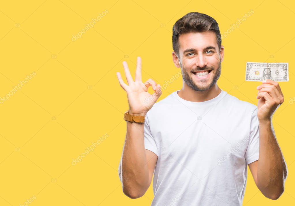 Young handsome man holding dollars over isolated background doing ok sign with fingers, excellent symbol