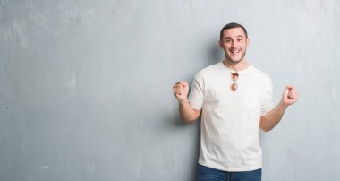 Beyaz delikanlı kutluyor güneş gözlüğü takıyor gri grunge duvarın üzerinden şaşırttı ve başarı için kaldırdı kollarını açarak hayret ve gözleri açık. Kazanan kavramı.