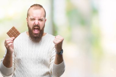 Genç hipster adam yeme çikolata izole üzerinde arka plan ile öfke, çılgın ve yükseltilmiş eliyle diye bağıran rahatsız ve hayal kırıklığına uğramış öfke kavramı bağırarak