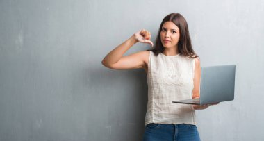 Grunge genç esmer kadın gri bilgisayar dizüstü kullanarak kızgın yüzü, eksi işareti aşağı antipati başparmak ile gösterilen, ret kavramı ile duvar