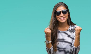 Genç güzel Arap kadın üzerinde güneş gözlüğü takıyor arka plan çok mutlu ve heyecanlı kaldırdı, silah ile kazanan jest yapıyor gülümseyen ve başarı için çığlık izole. Kutlama kavramı.