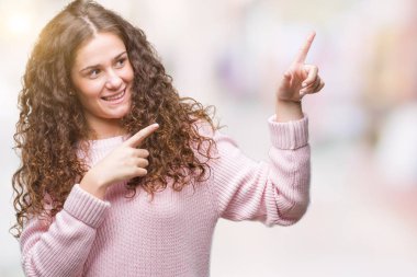 Güzel esmer kıvırcık saçlı Genç kız gülümseyerek ve iki elleri ve parmakları ile tarafına işaret kamera bakarak izole arka plan üzerinde pembe kış kazak giyiyor.