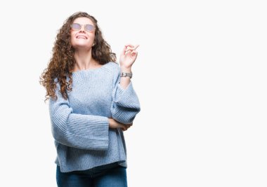 Kameraya bakarak yan el ve parmak ile işaret yüzünde kocaman bir gülümseme ile izole arka plan üzerinde güneş gözlüğü takıyor güzel esmer kıvırcık saçlı Genç kız.