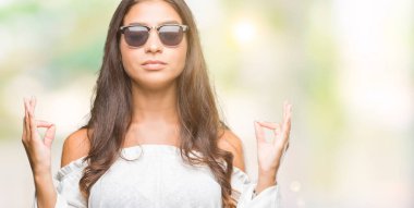 Genç güzel Arap kadın giyiyor güneş gözlüğü izole arka plan üzerinde dinlenmek ve gülümseyen gözlerle kapalı parmak jestle meditasyon yapıyor. Yoga kavramı.
