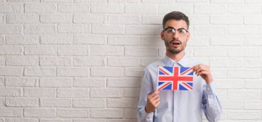 Genç yetişkin adam İngiltere bayrağı korkmuş şok bir sürpriz yüz, korkuyor ve heyecanlı korku ifadesi ile tutarak tuğla duvarın üzerinden
