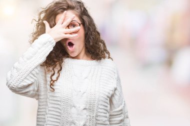 Mahcup ifadeyle parmaklarımın arasından seyir yüz ve gözler el ile kapsayan şok bakmak izole arka plan üzerinde kış kazak giyen güzel esmer kıvırcık saçlı Genç kız.