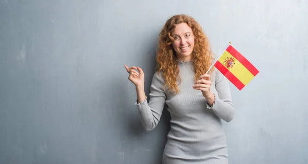 Młody Rude Kobiety Ponad Szary Tło Ściany Trzymając Flagę Hiszpanii — Zdjęcie stockowe