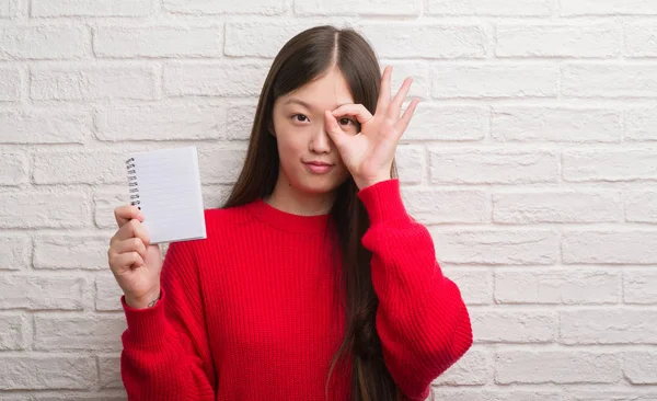 在砖墙上的年轻的中国妇女显示空白笔记本与愉快的脸微笑做 标志与手在眼睛看通过手指 — 图库照片