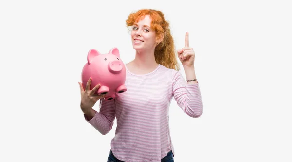 年轻的红头发的女人抱着小猪银行惊讶的想法或问题指着一个快乐的脸 — 图库照片