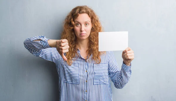 Young redhead woman over grey grunge wall holding blank card with angry face, negative sign showing dislike with thumbs down, rejection concept