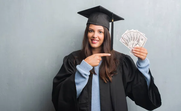 ドルを手と指で指している非常に満足して保持している大学院の制服を着て不潔な灰色の壁を越えて若いブルネットの女性 — ストック写真
