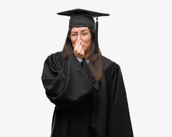 年轻的拉美裔妇女穿着毕业帽和制服闻到一些恶臭和恶心 无法忍受的气味 用手指在鼻子上呼吸 难闻的气味概念 — 图库照片
