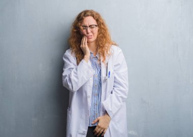Genç redhead doktor kadın diş ağrısı veya dişlerde dişlerle ilgili hastalık nedeniyle ağız acı ifadesi ile el ile dokunmak bir palto giyiyor gri grunge duvarın üzerinden. Diş hekimi kavramı.