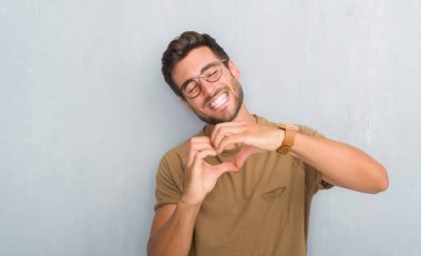 Yakışıklı delikanlı aşık kalp simgesi ve elleriyle şekil gösteren gülümseyen gözlüklü gri grunge duvarın üzerinden. Romantik kavramı.
